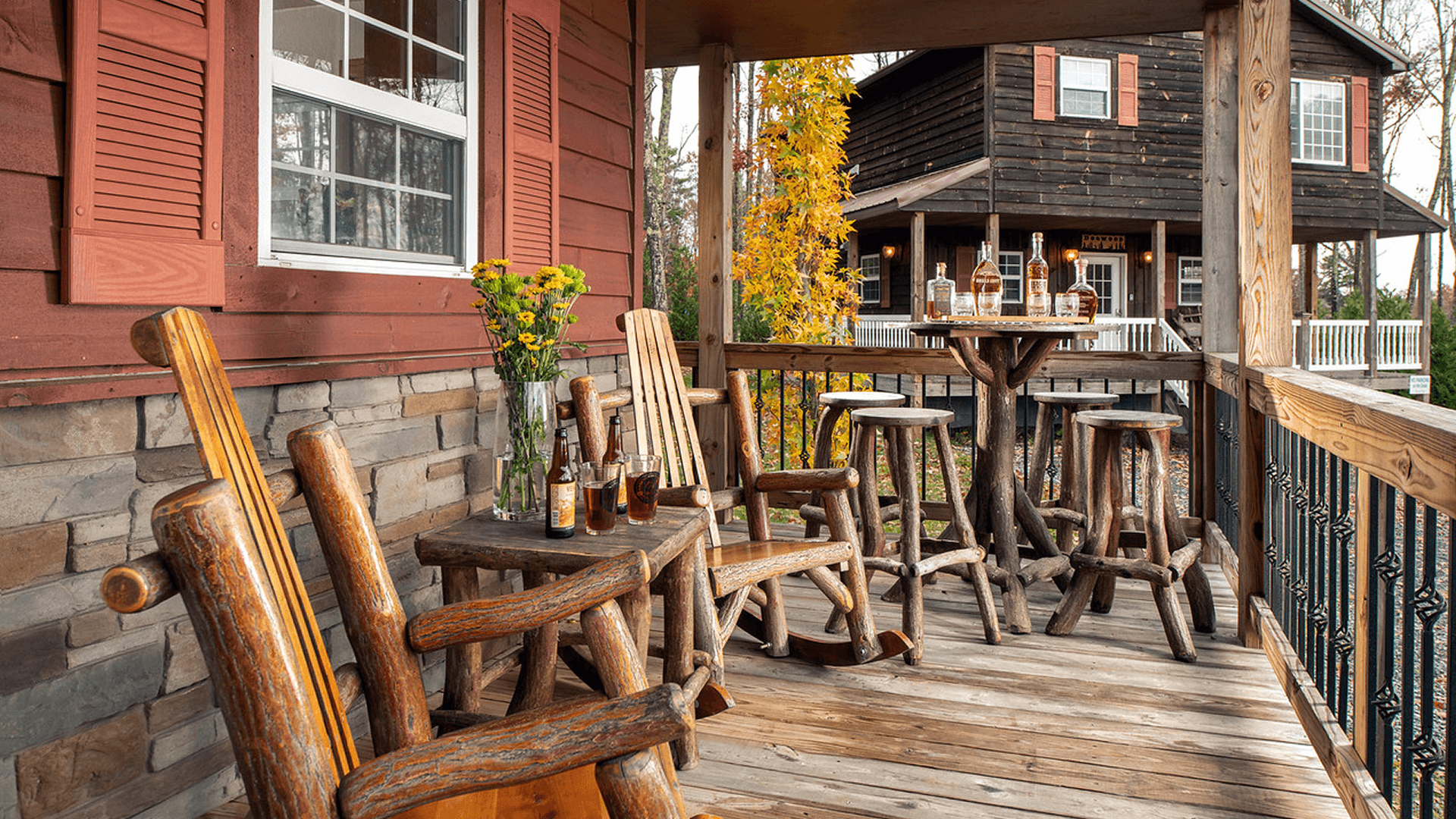 Front porch of chalet, heavy wood furniture, chairs, rockers, low and high tables, with drinks in glasses and another chalet in the background