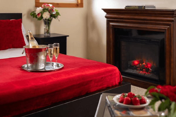A cozy bedroom featuring a red bedspread, champagne on a tray, and a fireplace with glowing embers.